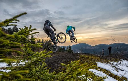 2 Mountainbiker beim Sprung auf Bikestrecke mit Schneeresten