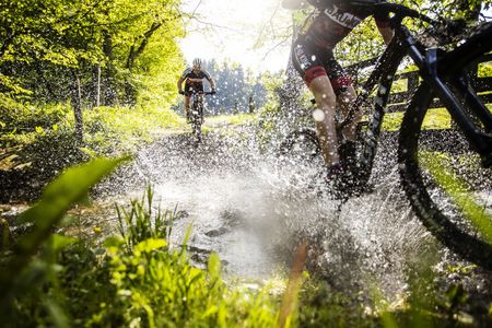 Ein Mann und ein Kind auf Mountainbikes fahren durch einen Bach, das Wasser spritzt in alle Richtungen